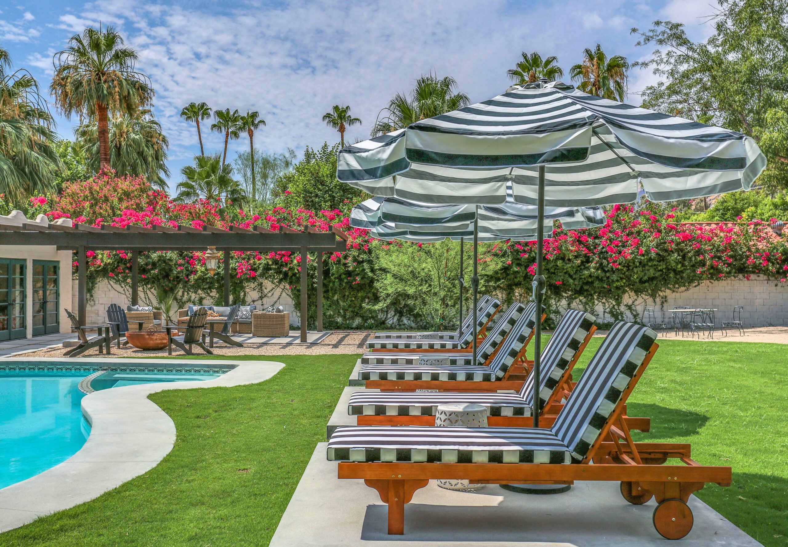 85 Four striped lounge chairs and umbrellas next to a pool, surrounded by green grass, trees, and pink flowers under a partly cloudy sky.